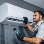 صورة خارجية فوق سطح بناية سكنية أو تجارية في منطقة الرقعي بالكويت، تظهر فني تكييف ماهراً يرتدي زي العمل ومعه حقيبة المعدات وهو يقوم بفحص مروحة التبريد الخارجية لوحدة تكييف مركزي (Central Package Unit). يظهر في الخلفية الطابع العمراني للمنطقة، مما يعكس سرعة الوصول والخدمة الفورية للسكان.